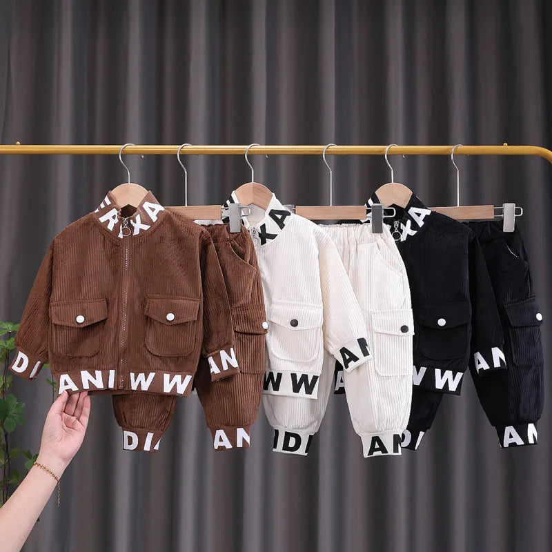 Children's clothing set with jackets and pants on hangers against a dark curtain background.