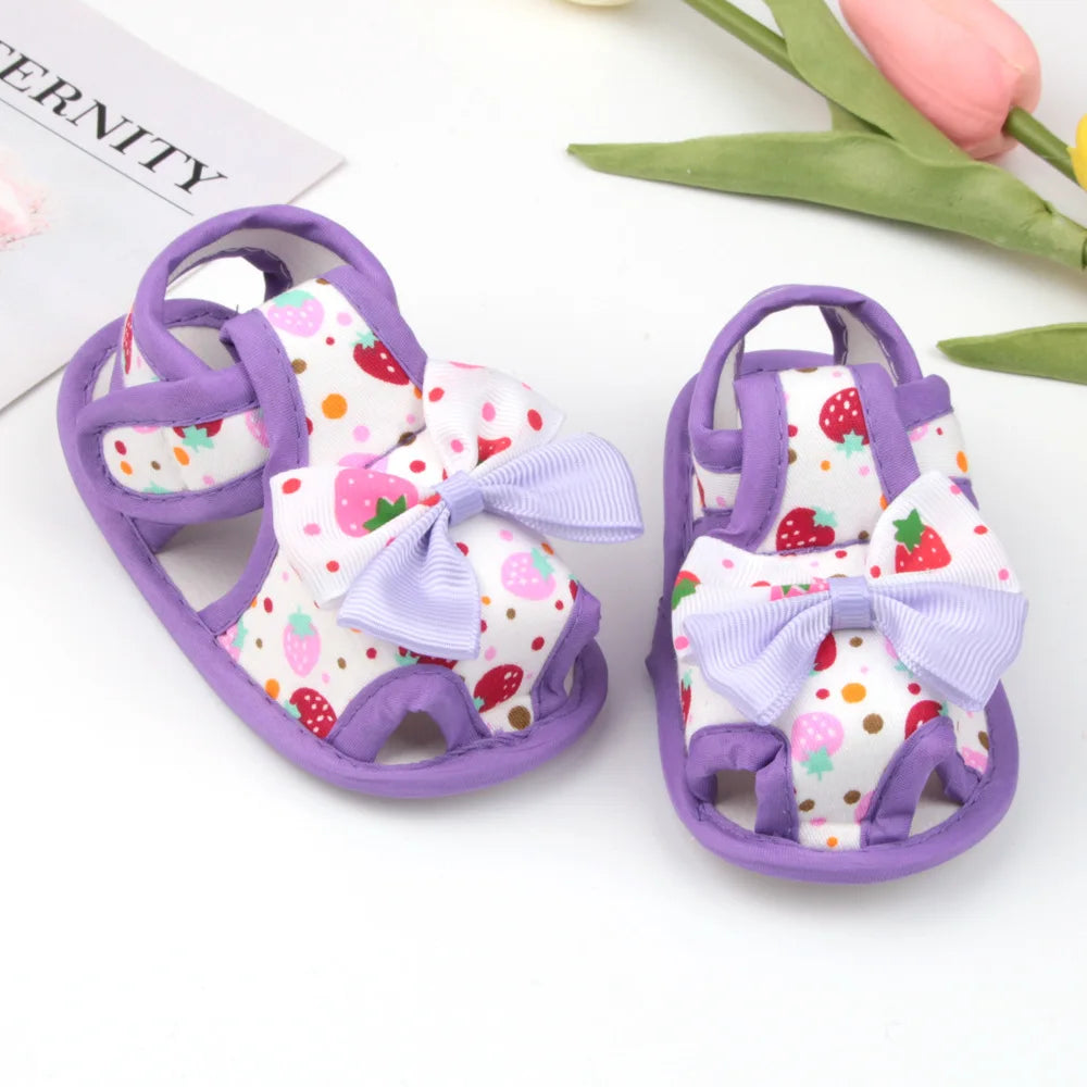 Pair of children's sandals with strawberry pattern and purple bows on a white background.