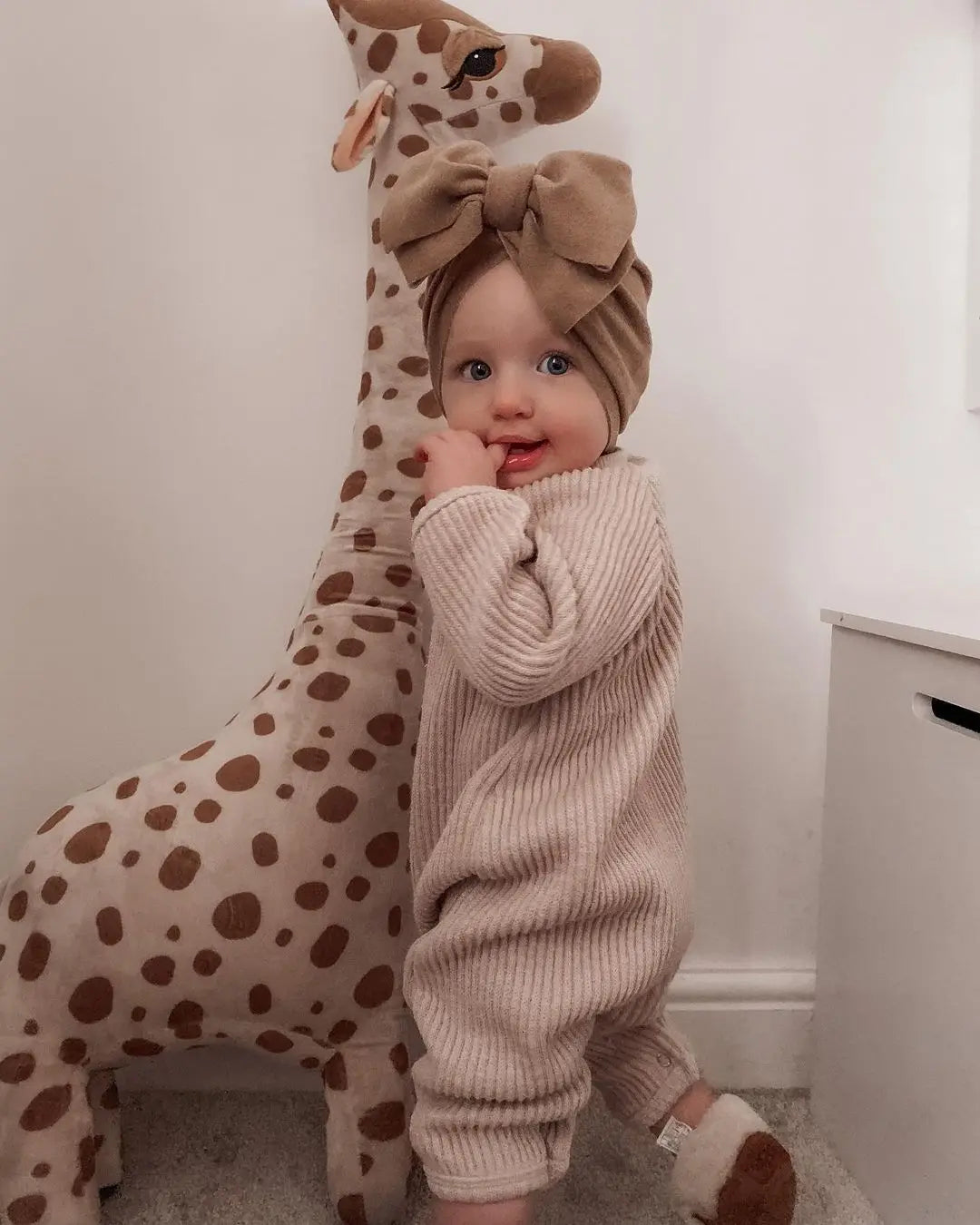 Baby in a ribbed sweater standing next to a plush giraffe toy indoors.