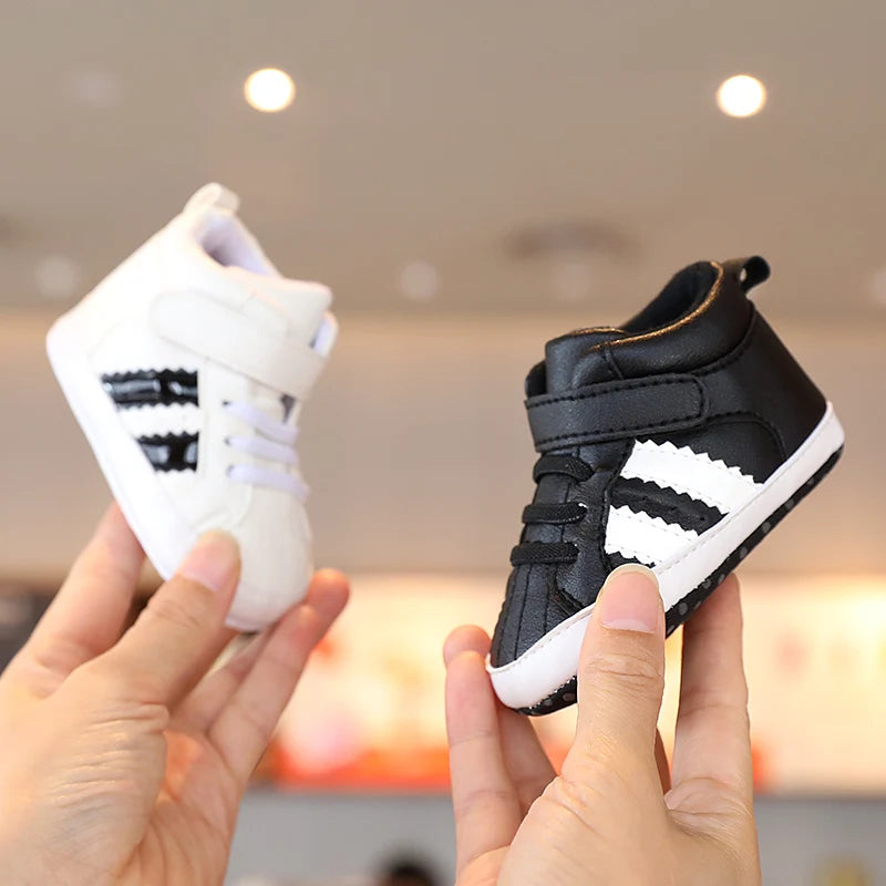 Two pairs of baby shoes, one white and one black with white stripes, held by a person against a blurred indoor background.
