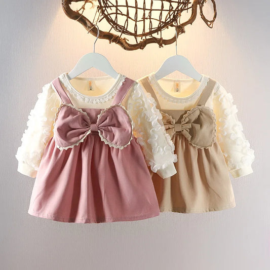 Two children's dresses with large bows on hangers against a neutral background