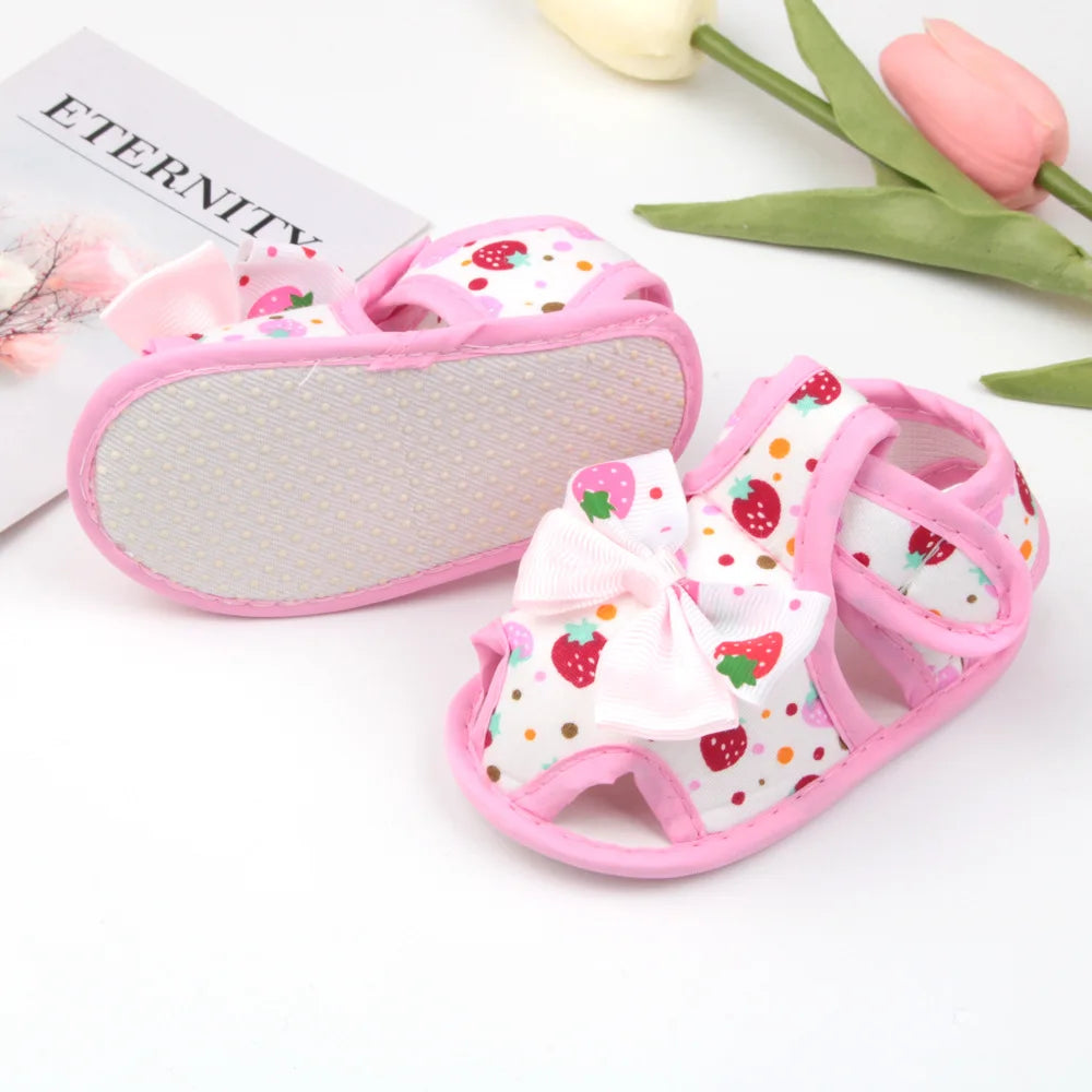 Baby sandal with strawberry pattern and pink bow next to a sanding block on a white background.