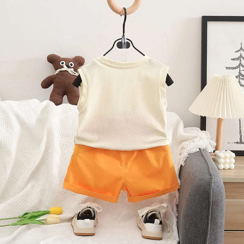 Children's outfit with white shirt and orange shorts on a hanger, with shoes and teddy bear in the background.