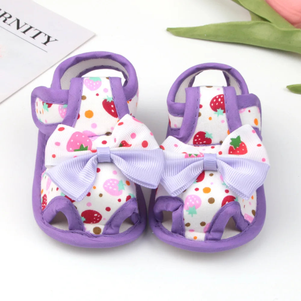 Pair of baby sandals with strawberry pattern and purple bows on a white background