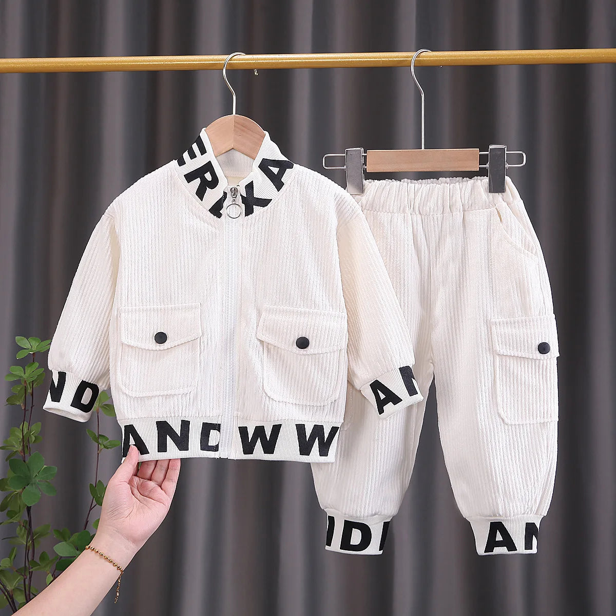 White children's jacket and pants with black lettering on hangers against a dark curtain background.