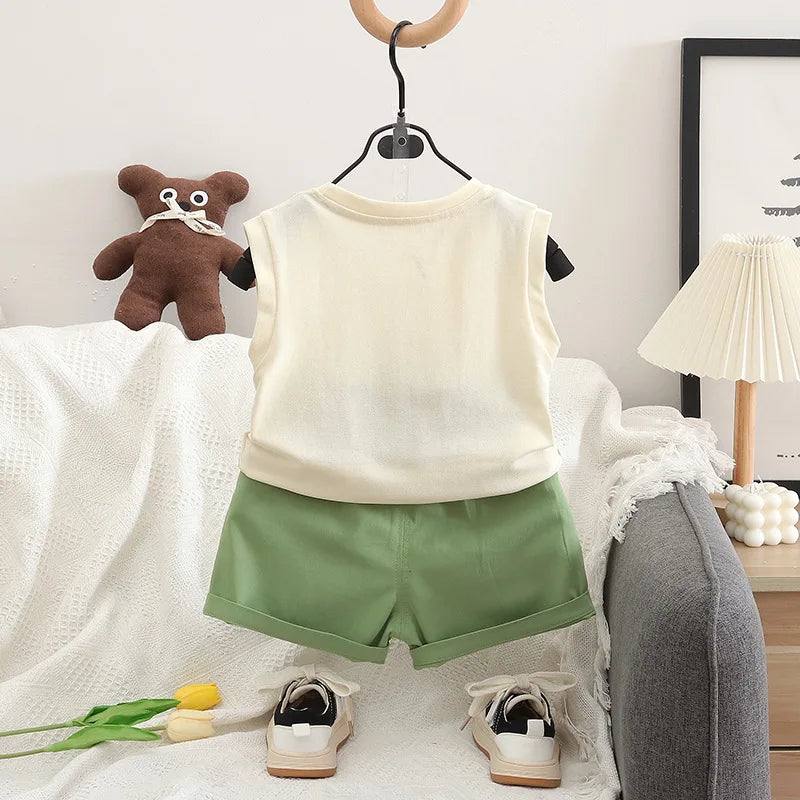 Children's outfit with beige sleeveless top and green shorts on a hanger, with shoes and a teddy bear on a bed.