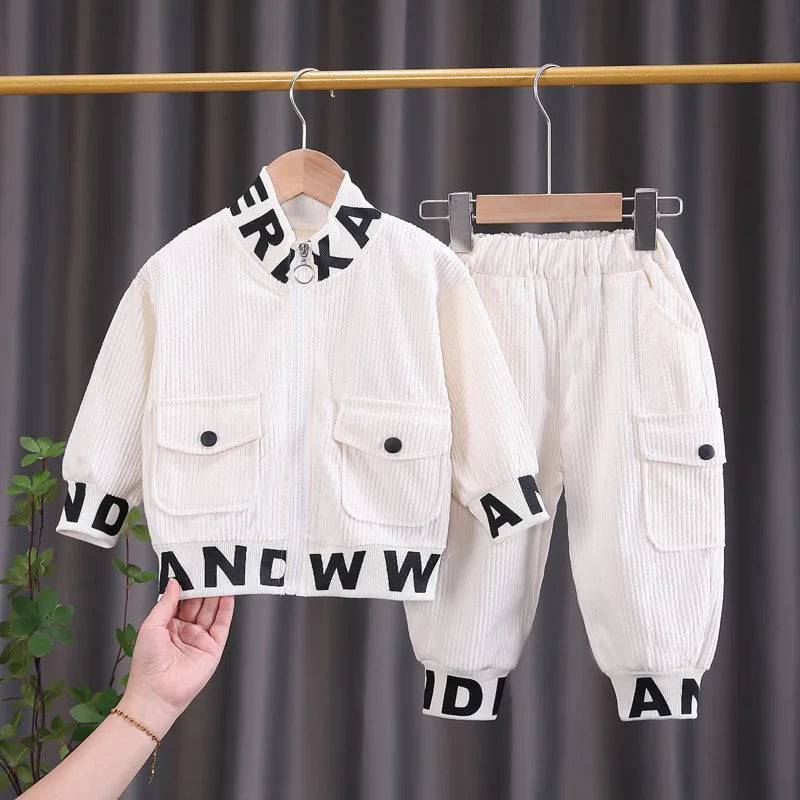 White children's jacket and pants with black lettering on hangers against a dark curtain background.