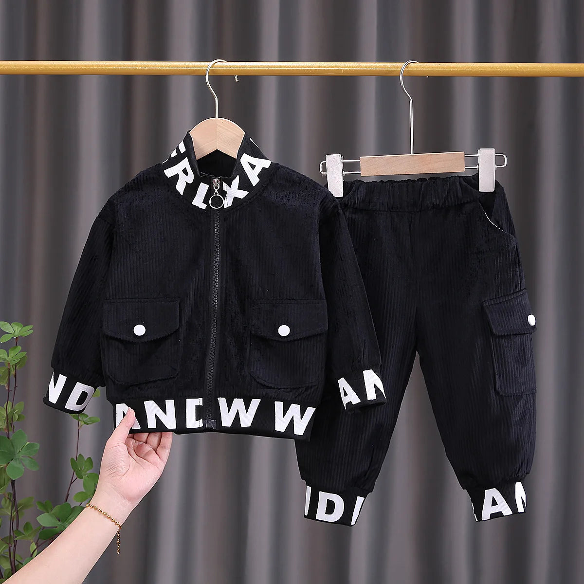 Black children's jacket and pants set with white text on hangers against a gray curtain background.